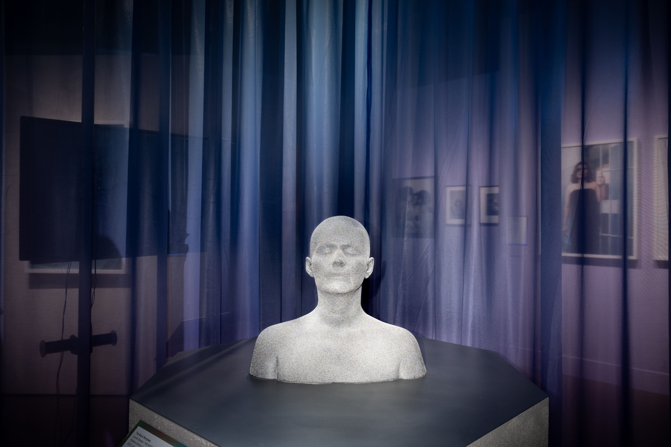A grey stone bust of the head and shoulders of a young male figure with eyes closed and no hair sits on an octagonal plinth. Behind it is a semi-transparent curtain throgh which can be seen a wider exhibition space with images mounted on walls.