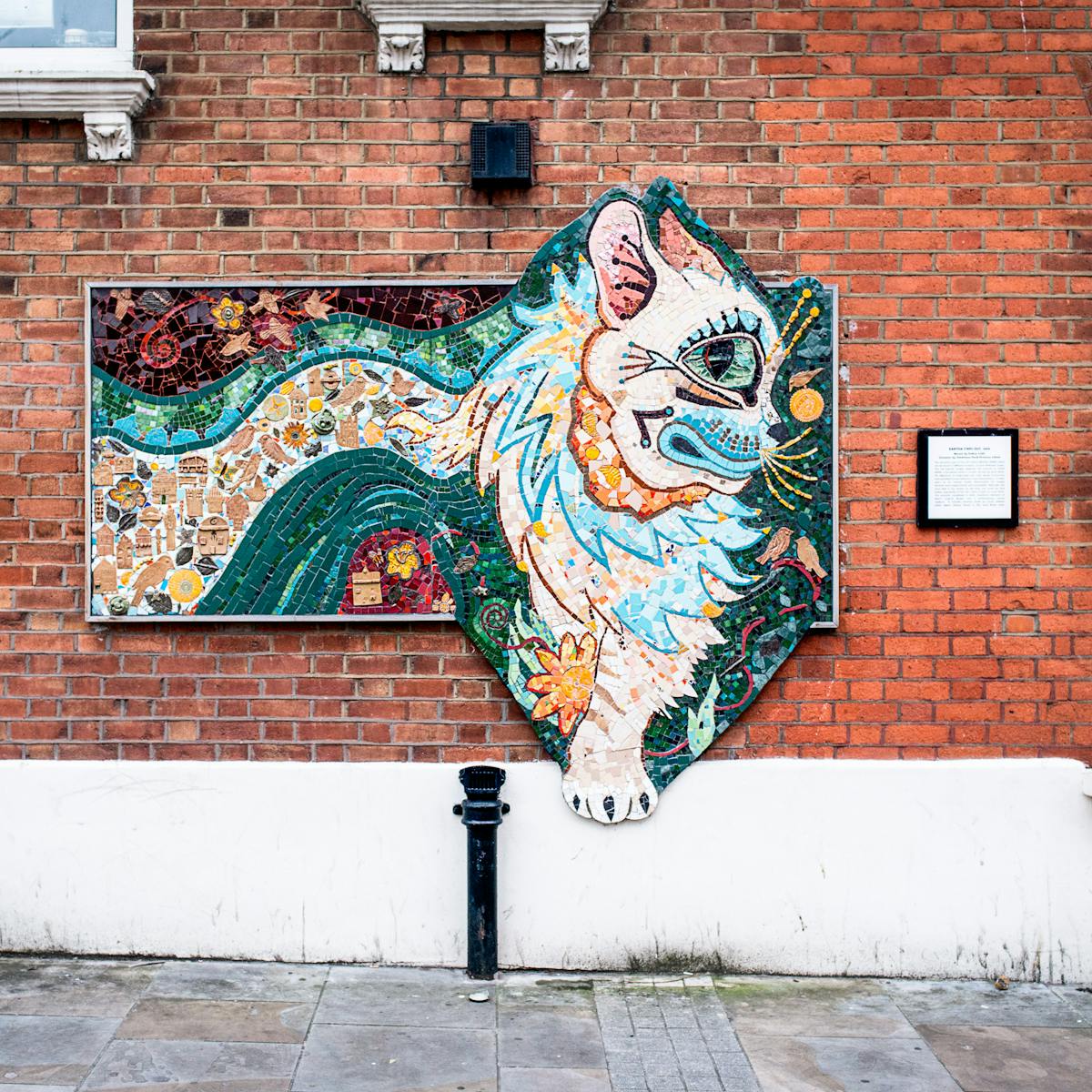 Photograph showing a brightly coloured abstract ceramic mosaic of a cat, in the style of Louis Wain. The mosaic is hung on the red brick wall of the outside of a property. Below the mosaic is the pavement and elements of street furniture. To either side are parts of window and an information plaque. 