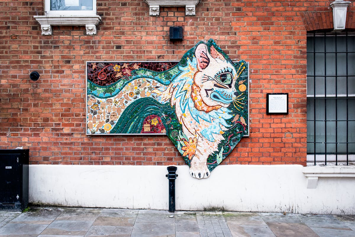 Photograph showing a brightly coloured abstract ceramic mosaic of a cat, in the style of Louis Wain. The mosaic is hung on the red brick wall of the outside of a property. Below the mosaic is the pavement and elements of street furniture. To either side are parts of window and an information plaque. 