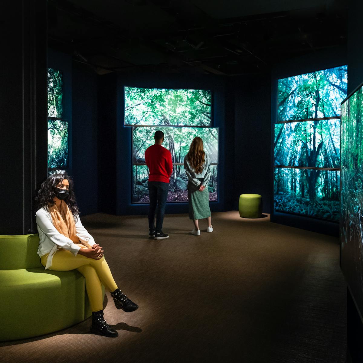 Photograph of a dark exhibition gallery space showing 4 separate large free-standing walls on which photographic prints of large forest scenes are displayed, filling the entire walls. The prints show tall tree trunks, green leaf canopy cover and ground level ferns. To the left of the gallery view is a circular padded green seat which wraps around a vertical iron girder. Sitting cross legged on the seat is a woman with long black hair, a white blouse and yellow leggings. She has an orange shawl around her neck and is wearing a black face covering. She is spotlit from a light almost directly above her. In the distance are two more visitors with their backs to the camera, standing looking at one of the forest scenes. To their right is a small green padded stool.