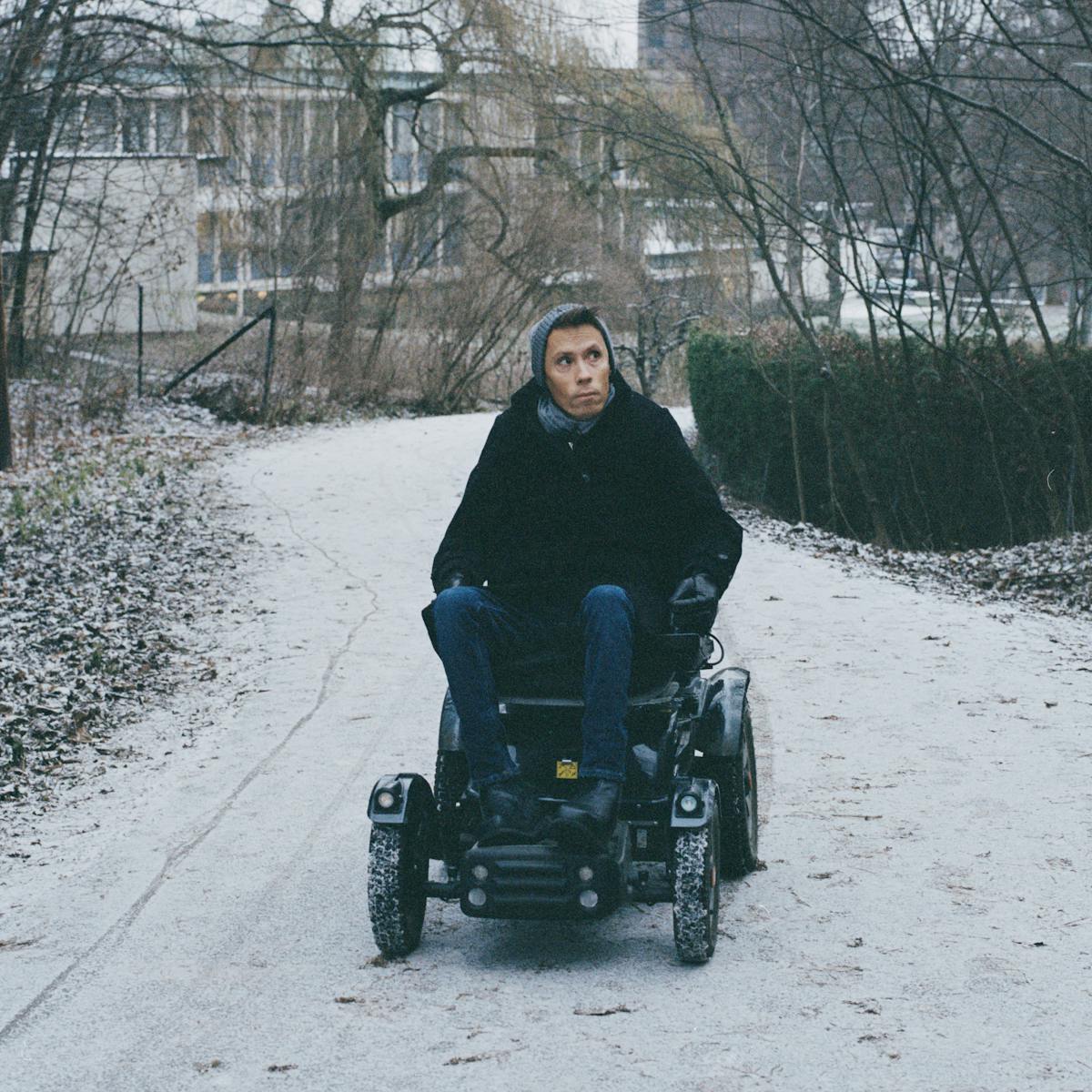 Photograph of a man using an off-road electric wheelchair. He is wearing jeans, a dark coat and a grey wooly hat and scarf. He is pictured driving along a snowy path, winding though winter woodland. Behind him in the distance are buildings. He is looking off to the right as he drives towards the camera.