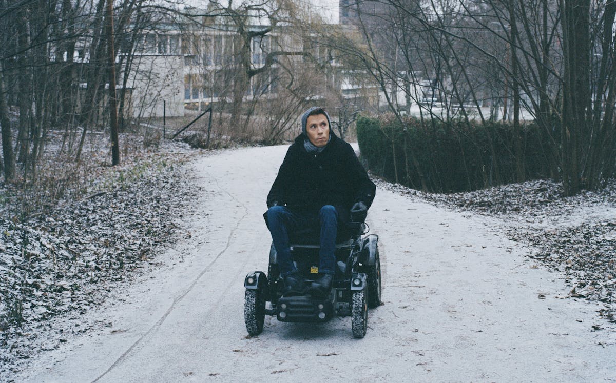 Photograph of a man using an off-road electric wheelchair. He is wearing jeans, a dark coat and a grey wooly hat and scarf. He is pictured driving along a snowy path, winding though winter woodland. Behind him in the distance are buildings. He is looking off to the right as he drives towards the camera.