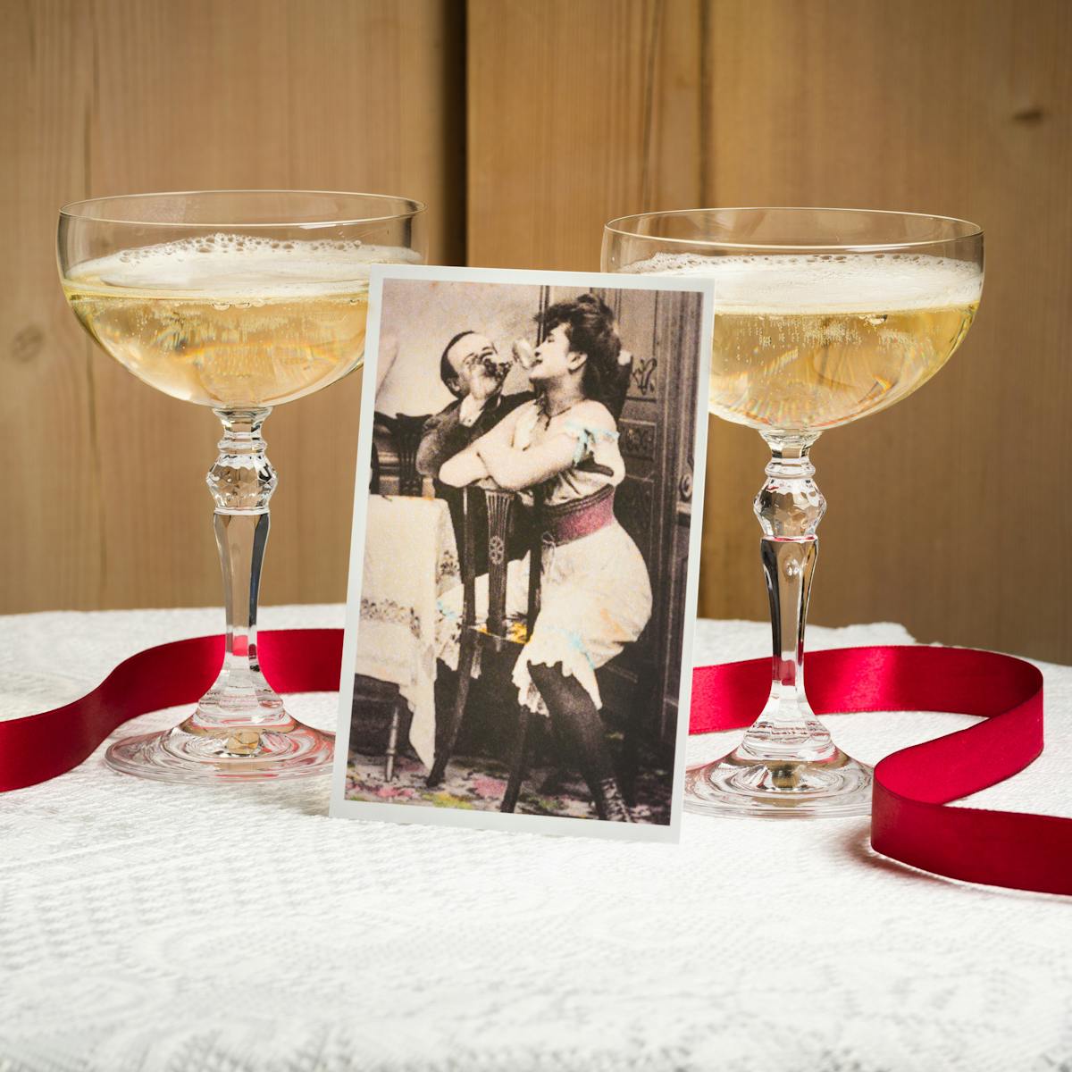 Colour photograph of a round table top covered in a white lace table cloth against a wooden panelled wall. On the table are two champagne coupes filled with sparkling champagne. Propped up against one of the glasses is a old hand tinted photographic print. The print is from the turn of the 20th century and show a woman in under garments sitting on a dining chair backwards, legs astride the backrest. Behind her a man is seated pouring a drink from a champagne glass into her mouth. A red ribbon snakes on its side around the bases of the glasses and the photographic print and off the side of the table.