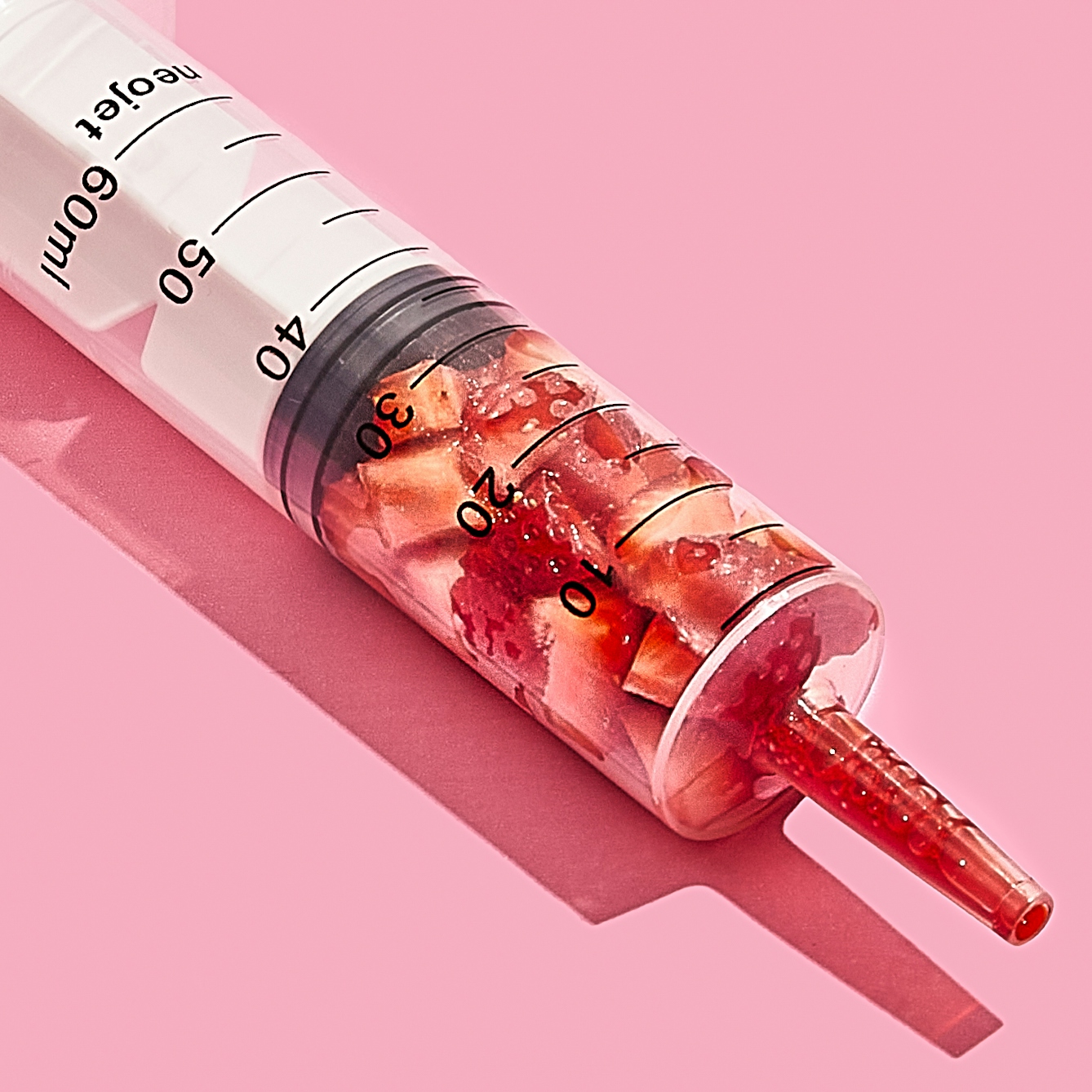 Photograph of a medical syringe lying on its side on a bright pink background. The syringe is full of chunks of cut up strawberry, compressed down into the lower half of the graduated tube. The nozzle of the syringe contains bright red strawberry juice. 