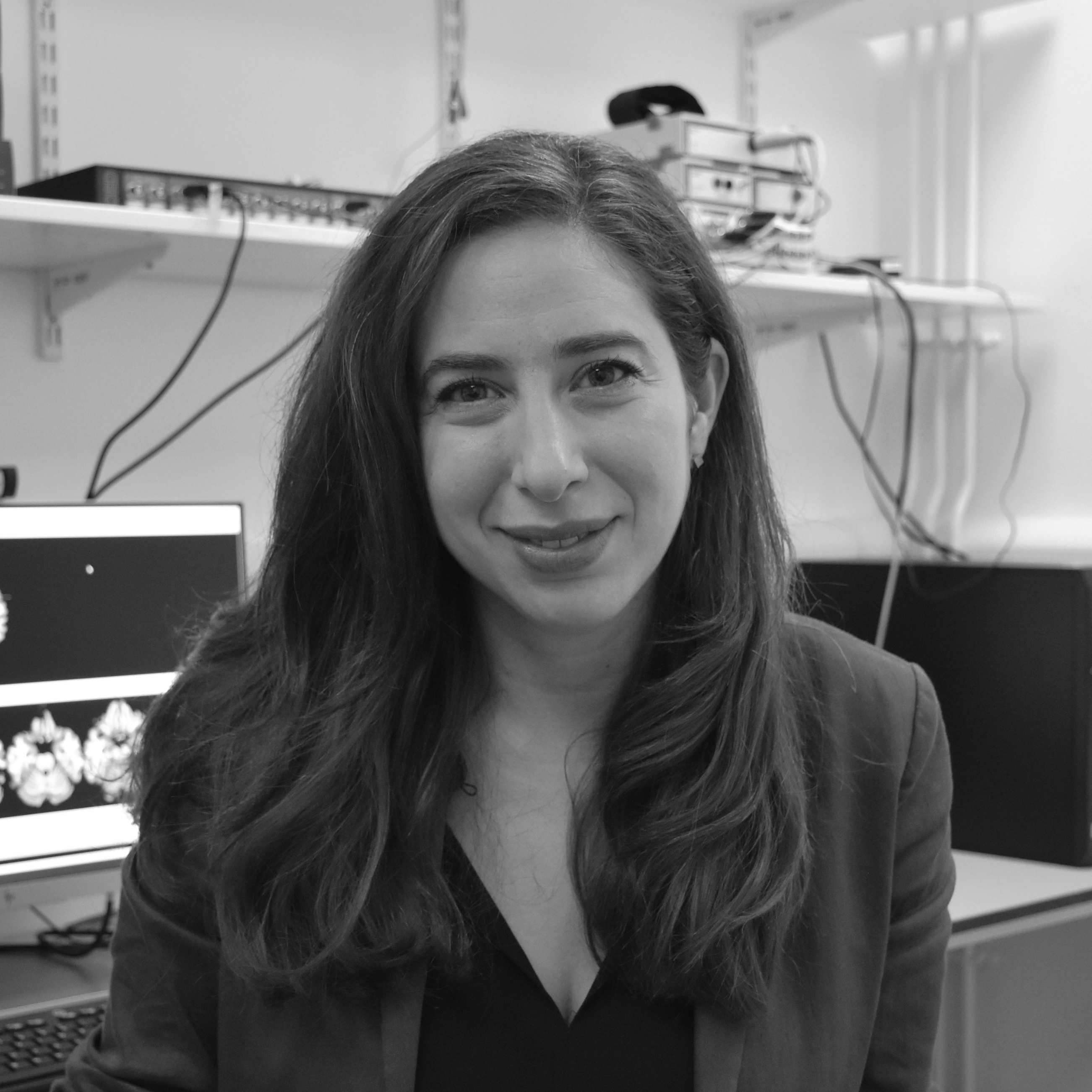 Black and white, head and shoulder photograph of a woman with long dark hair. She is wearing a dark top and jacket. In the background are shelves and a desk with a computer.