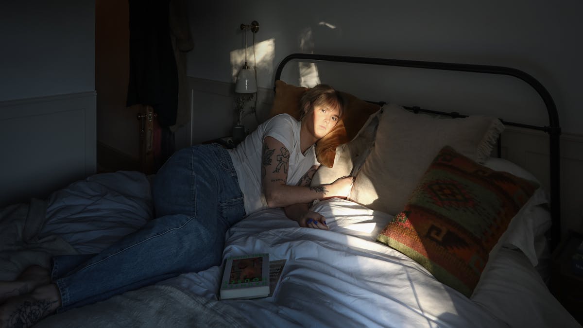 Photograph of a young woman lying on her side on a double bed, in a bedroom. She is wearing blue jeans and a white t-shirt. Her face is lit up in a shaft of sunlight which travels diagonally across the bedclothes and over her face and the wall behind. Her eyes are open and she is looking past the camera into the distance. On her arms are several tattoos and a couple of closed book rest on the bed next to her.