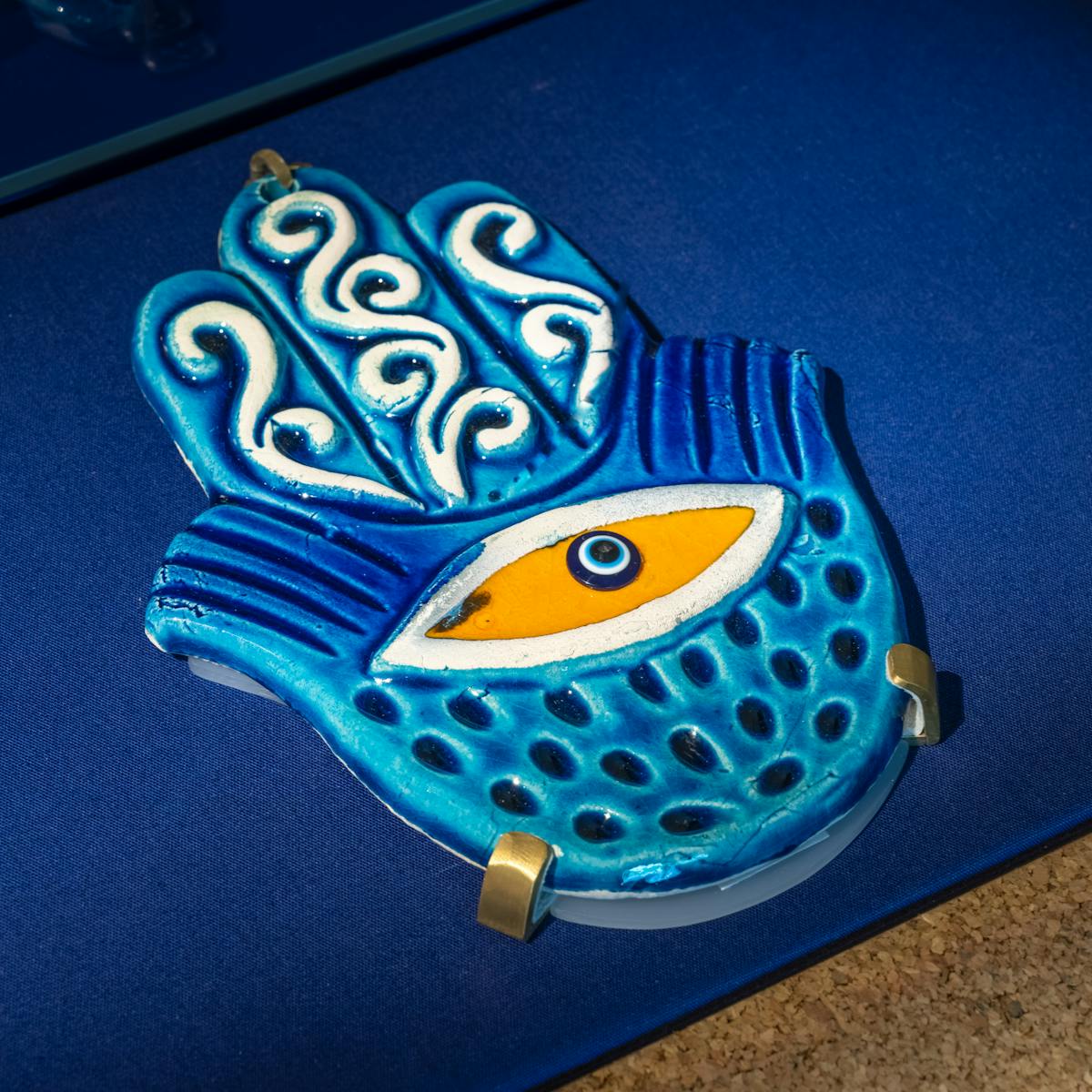 Vivid blue ceramic hand-shaped charm with a bright yellow eye in its palm. The surface of the hand is decorated with patterns of swirls, stripes and dots. 