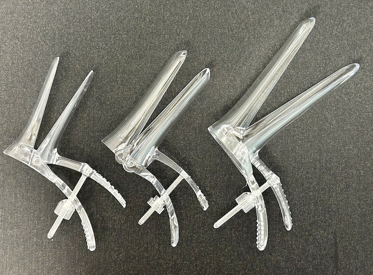 Three sizes of clear plastic disposal vaginal speculums. The three devices lie in a row in increasing size, all in the open position.