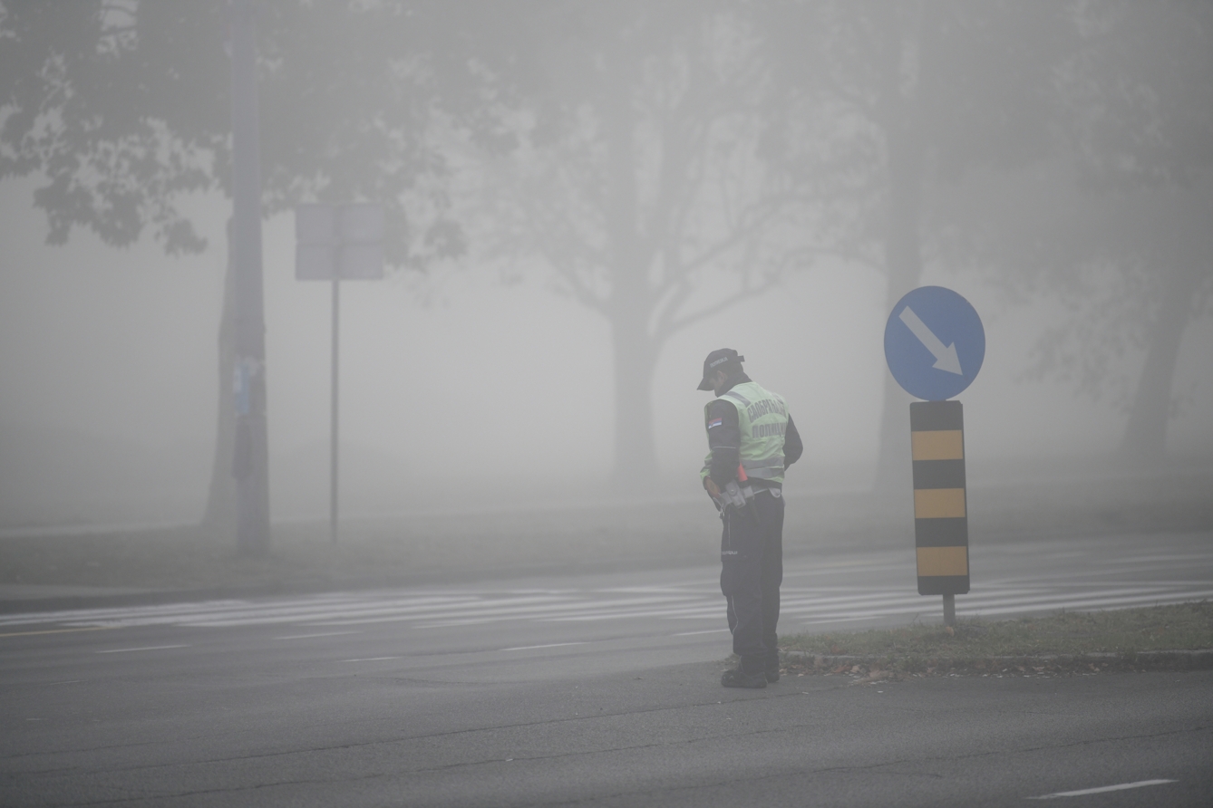 Photograph showing the intersection of two streets, with a pedestrian crossing in the middle. It is very misty, and the mist is enveloping a line of several leafy trees and a road sign in the background. There is a man in a hgih viz vest next to a road sign in the foreground. 