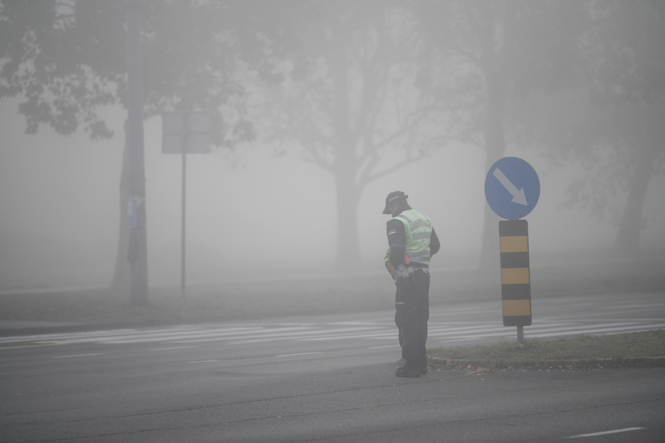 Photograph showing the intersection of two streets, with a pedestrian crossing in the middle. It is very misty, and the mist is enveloping a line of several leafy trees and a road sign in the background. There is a man in a hgih viz vest next to a road sign in the foreground.