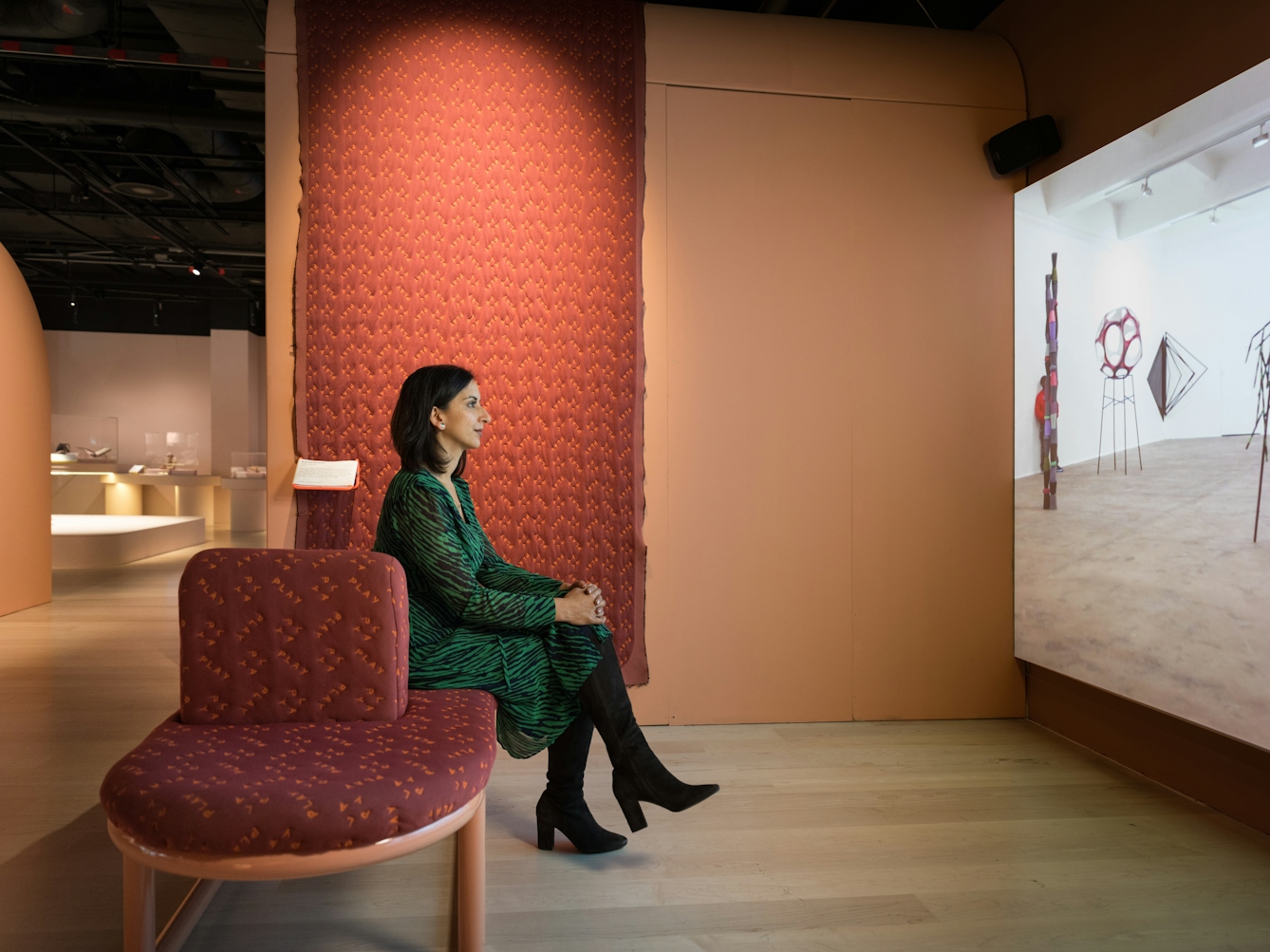 Photograph of a woman in an exhibition gallery space sitting on an upholstered bench, with her hand clasped around her crossed legs. She is watching a large projection screen to the right of frame.