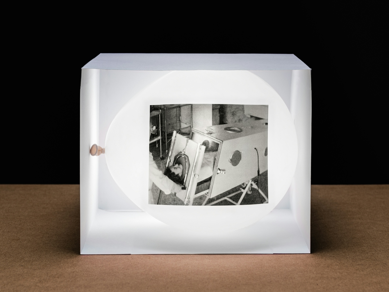 Photograph of a white inflated balloon lying horizontally within a box made of white card which is open on the side facing the camera. The balloon and box are resting on a wooden tabletop with a horizon line against a black background. The balloon looks like it is illuminated from within. On the side of the balloon is a rectangular, monochrome archive film still. The still shows a woman lying in a large metal box with her head protruding from one end. She is in the process of being slid into the box by someone’s arms entering the frame from the left. The contraction is an iron lung.