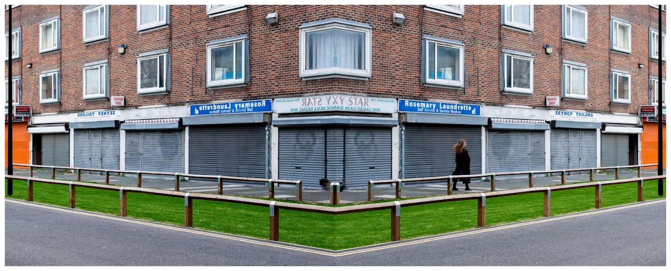 Photographic panorama showing a small high street scene with rows of shops with their shutters closed. The panorama is mirrored down its vertical centre line except that a small human figure walking down the street appears in the right half, but not in the left half.