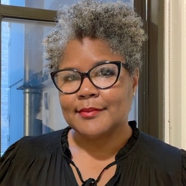 Headshot of a person with short, curly grey hair, dark-framed glasses and a black top.