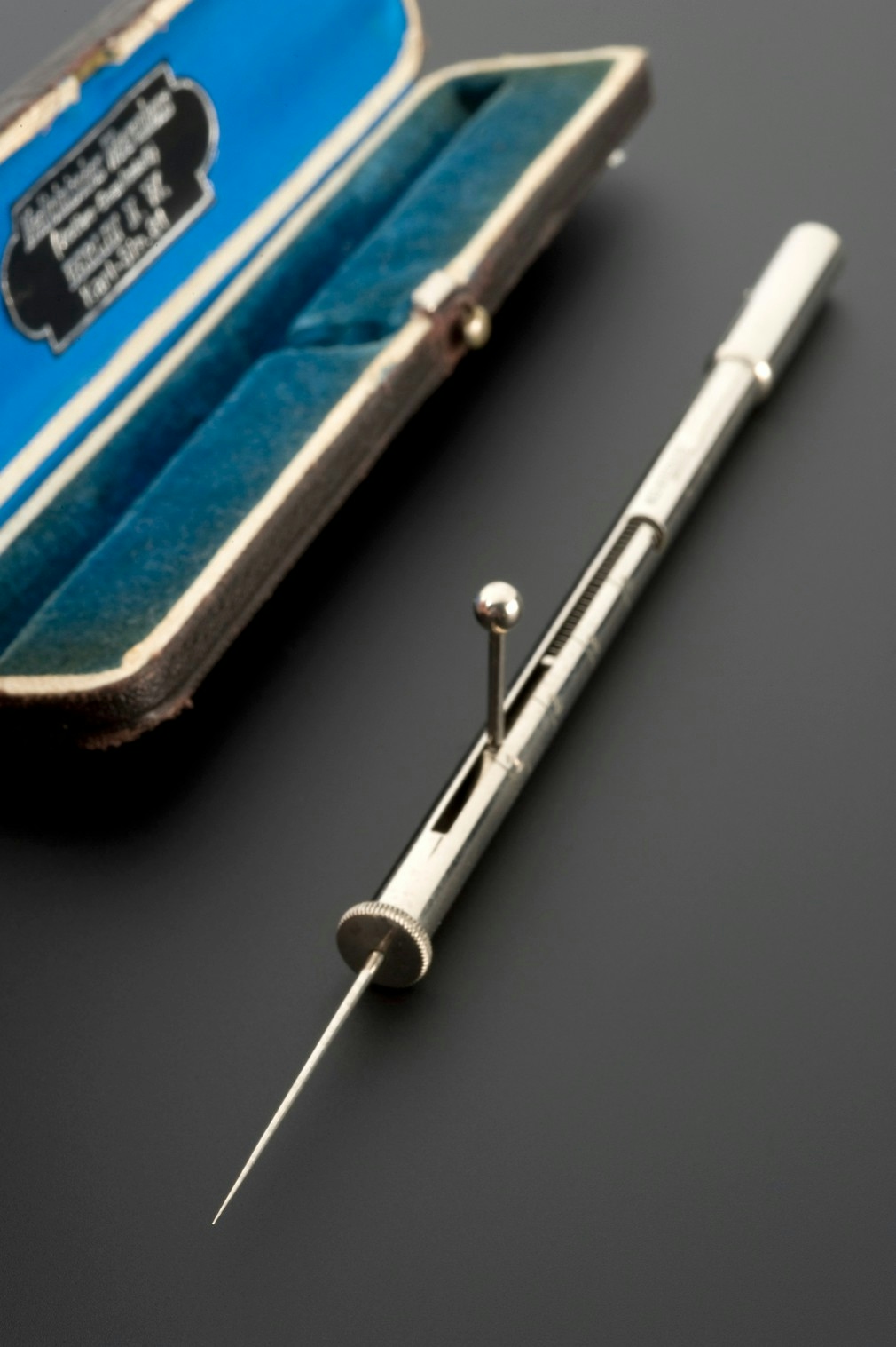 Photograph of an instrument case (in the background, lined with blue velvet) and a silver instrument called an algesimeter, which is a needle attached to a tube with a measure on the side to record the depth that a person is pricked before pain is expressed.
