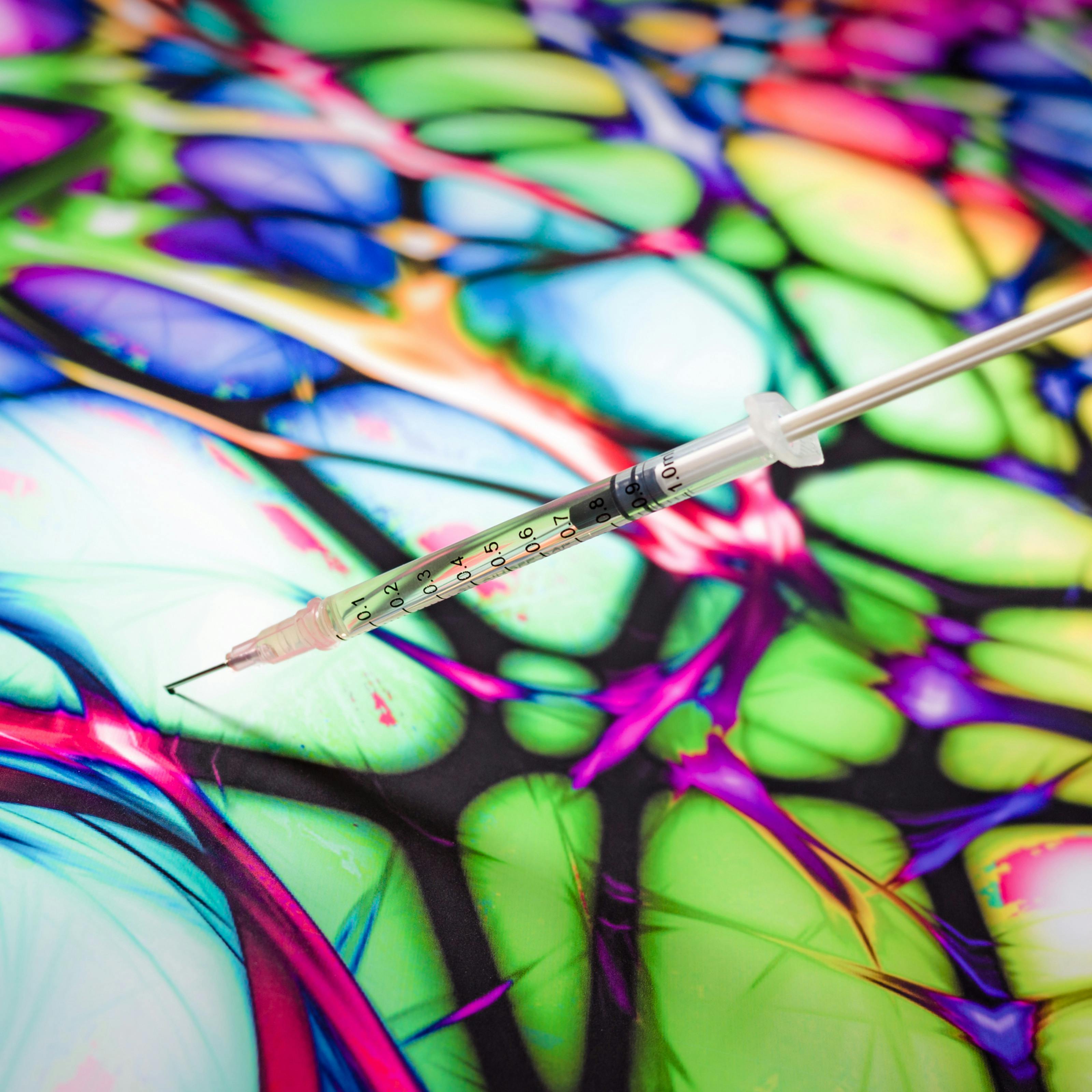 Photograph of syringe filled with liquid sticking out of a colourful background.