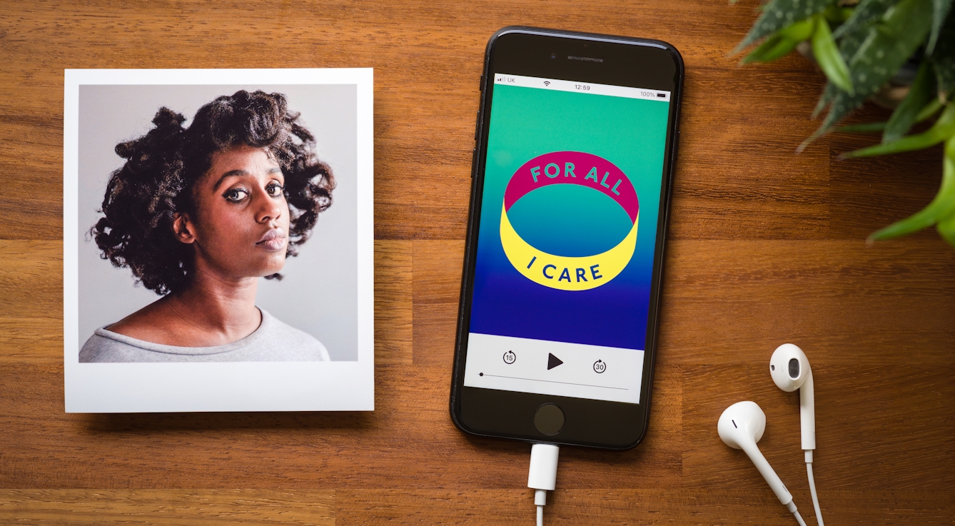 Photograph of a smartphone on a wooden tabletop. Plugged into the phone is a pair of white earbud headphones. On the screen of the phone is a podcast screen with a graphic identity of a coloured ring with the words 'for all I care' written around the ring,. The colours are green, blue, yellow and magenta. To the top right of the image is the green of a pot plant. To the left of the image is a photographic print of a portrait of a woman in a grey top against a grey background, she is looking to camera.