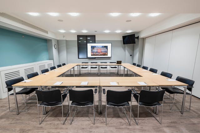Photograph of the Steel room at the Wellcome Collection. 

Photograph shows a boardroom set-up, with desks laid together in a rectangle shape with chairs around the outside, and a presenting screen at the front of the room. 