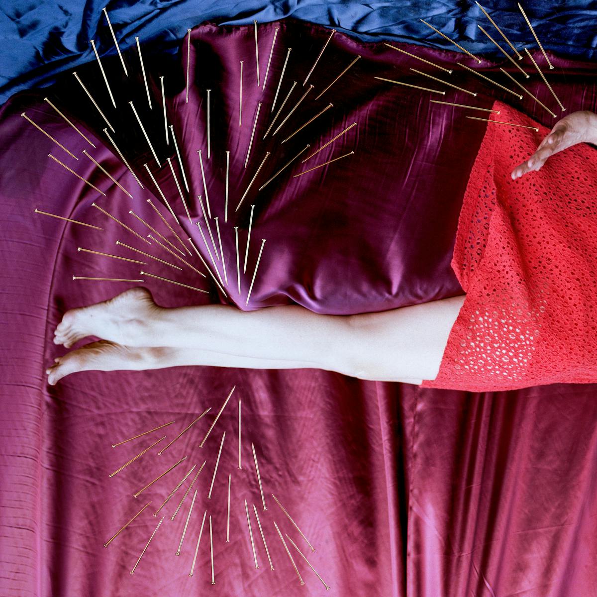 Artwork created with a colour photographic print of a female figure in a bright red dress, set against a purple and blue draped silk background. She is lying horizontally in the frame, face down, caught as if in mid fall.  Her hair and dress are blown upwards, toes and fingers outstretched. Her face is obscured by her hair. Her body is surrounded by groups of dress pins, laid on top of the photographic print. The pins are arranged as if they are a flight of arrows directed at her body. One group attacks her legs from underneath, another from above. A separate group flies towards her neck and chest.