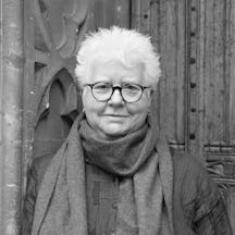 Black and white head and shoulders portrait of Val McDermid who has short white hair and is wearing round glasses, a coat and a scarf.