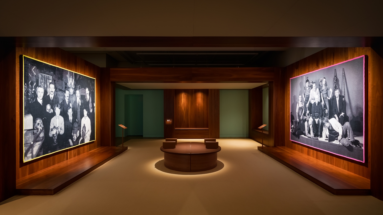 Photograph of a gallery exhibition space showing two large tapestries hung on opposite wooden walls, facing one another. The tapestries are made up of two black and white, archive photographs, one from the early 20th century and the other from the late 20th century. Both show a group of white men and women gathered in a group holding museum artefacts in their hands or on their laps. The tapestry on the left has a yellow border and the one one the right a pink border. Surrounding the tapestries is a wider wooden structure containing seating and alcoves. The hues of the scene are warm browns and olive greens.