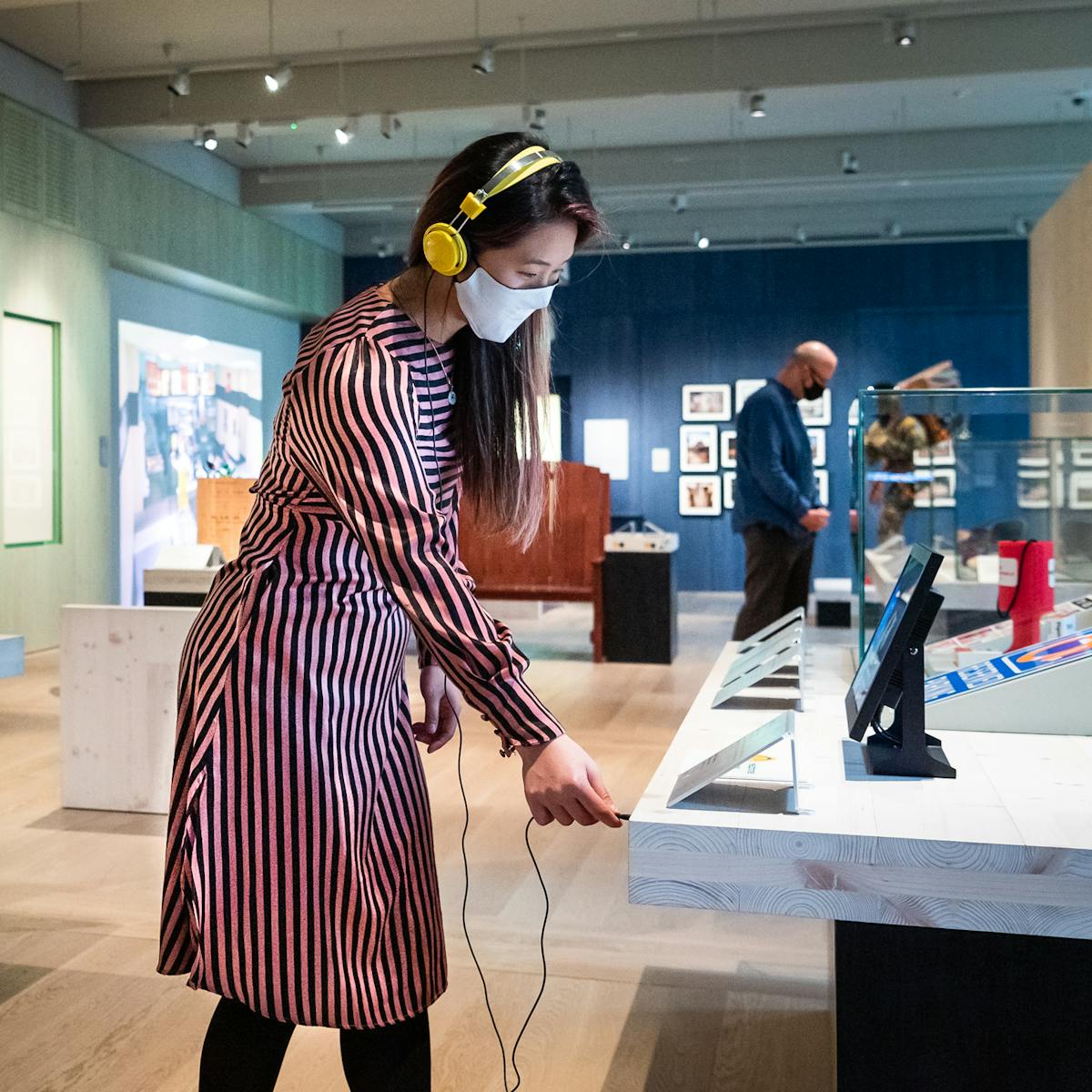 Photograph of a museum gallery space with display cases and exhibits. In the foreground is a woman wearing a face covering and a pair of yellow over the ear headphones. She is in the process of plugging the headphones into the socket of an audio exhibit. To the right of her is another woman also wearing a face covering who is looking up at a transparent model of human being. In the far distance is a man, also wearing a face covering who is exploring the exhibiton.