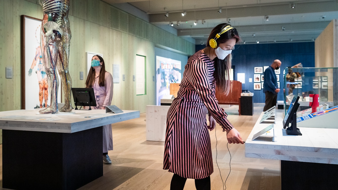 Photograph of a museum gallery space with display cases and exhibits. In the foreground is a woman wearing a face covering and a pair of yellow over the ear headphones. She is in the process of plugging the headphones into the socket of an audio exhibit. To the right of her is another woman also wearing a face covering who is looking up at a transparent model of human being. In the far distance is a man, also wearing a face covering who is exploring the exhibiton.