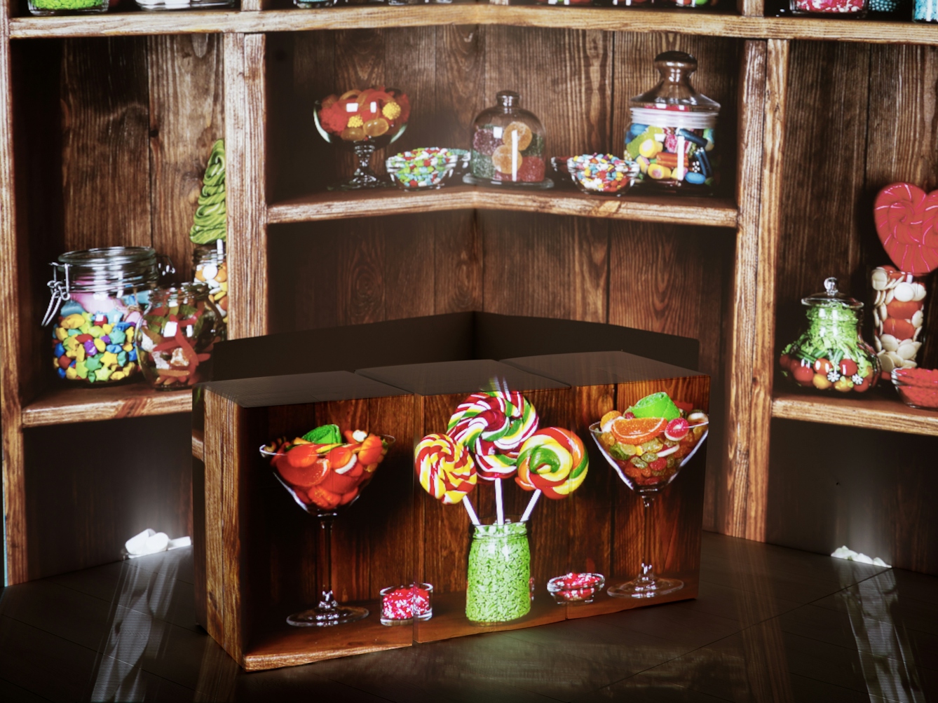 Photograph of the corner of a room showing the walls and a section of the wooden floor. On the floor is an arrangement of cardboard boxes. Onto the whole scene is projected an image of a sweet shop shelving with jars of sweets. The boxes have assumed the shape of the shelves and have jars of sweets projected onto their sides.