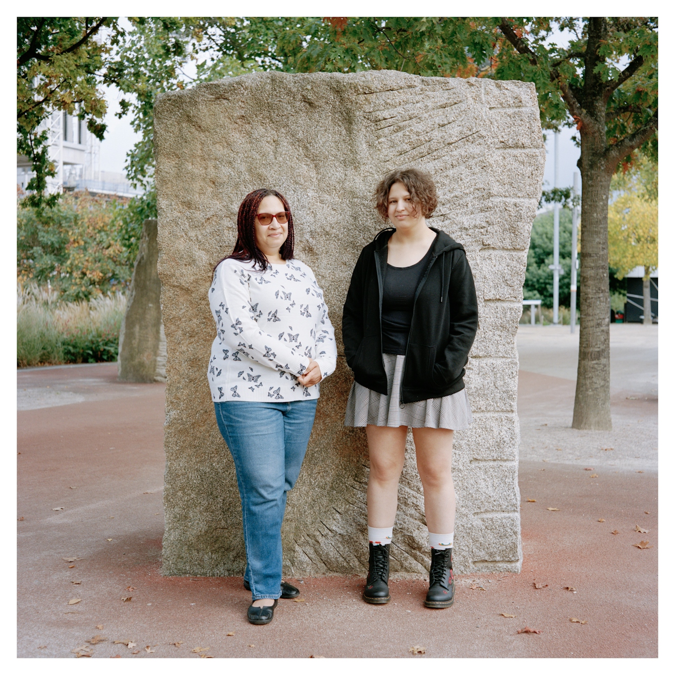 Full-length portrait photograph of two women, Amanda and Sophie, standing against a large rock in a park.  

The woman on the left has long braided hair and is wearing sunglasses, a white patterned top and blue jeans. She is looking at the camera and is smiling slightly. 

The woman on the right has short curly hair and is wearing a black zipped jumper, black t-shirt, grey short skirt and boots. She is also looking at the camera with a slight smile. 

Behind them there are several trees and plants in the park, the ground is tarmac. 