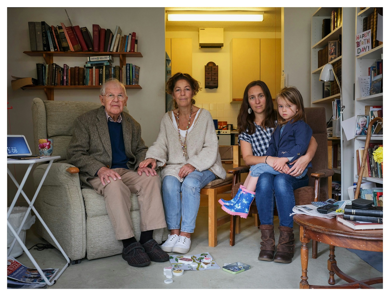 Family group photograph showing four generations of the same family. The great-grandfather sits on the left in a large armchair, by his side sits the grandmother whose hand rests on his. Next to her sits the grandmother who rests her hand on the knee of the granddaughter who is holding the great-granddaughter on her knee. They are all seated on chairs in a living room, surrounded by bookshelves and furniture. They all look straight into the camera.