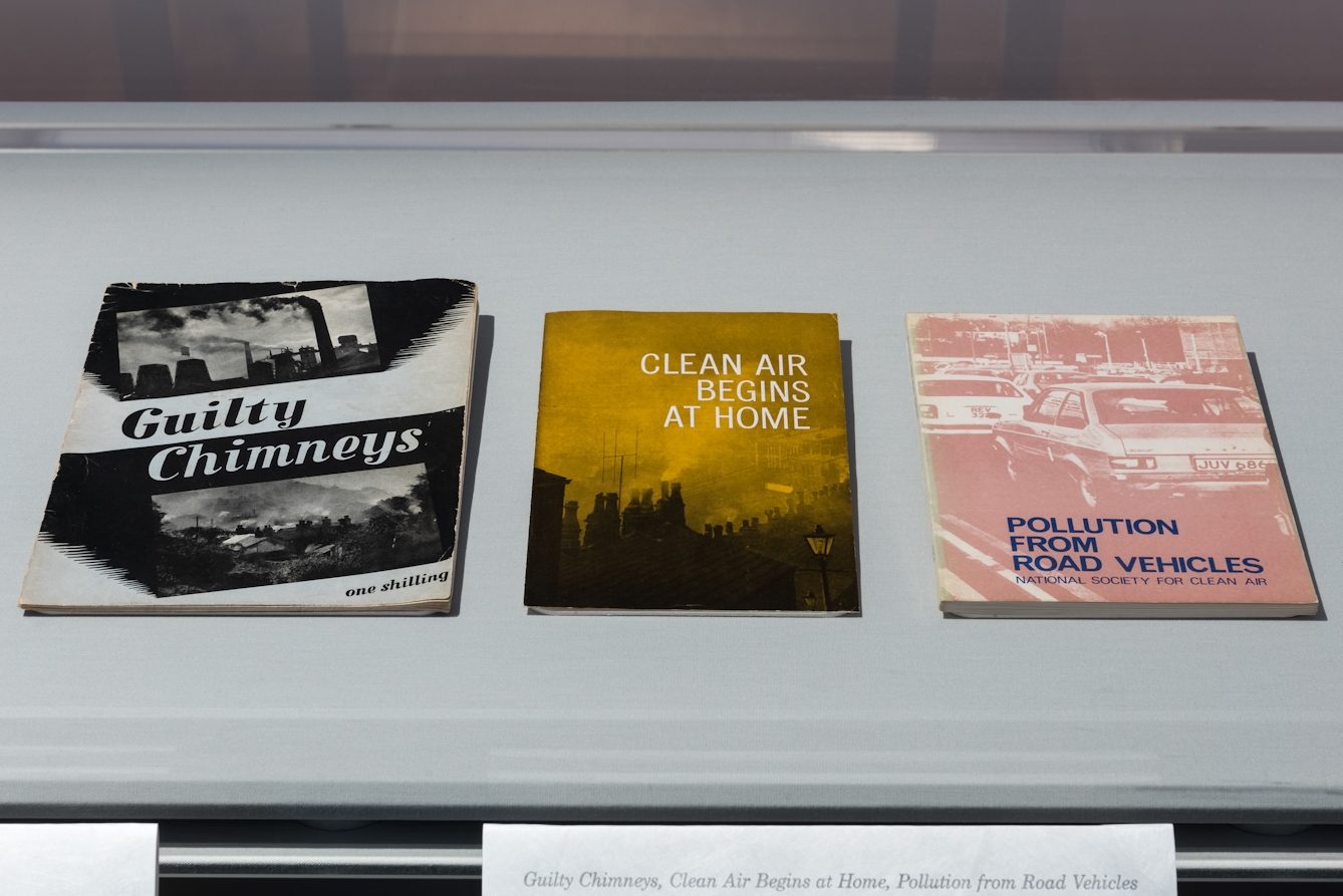 Colour photograph of three books laid next to eachother on a flat white surface. The book on the left is titled 'Guilty Chimneys'. The book in the centre is titled 'Clean air begins at home'. The book on the right is titled 'Pollution from road vehicles'. 