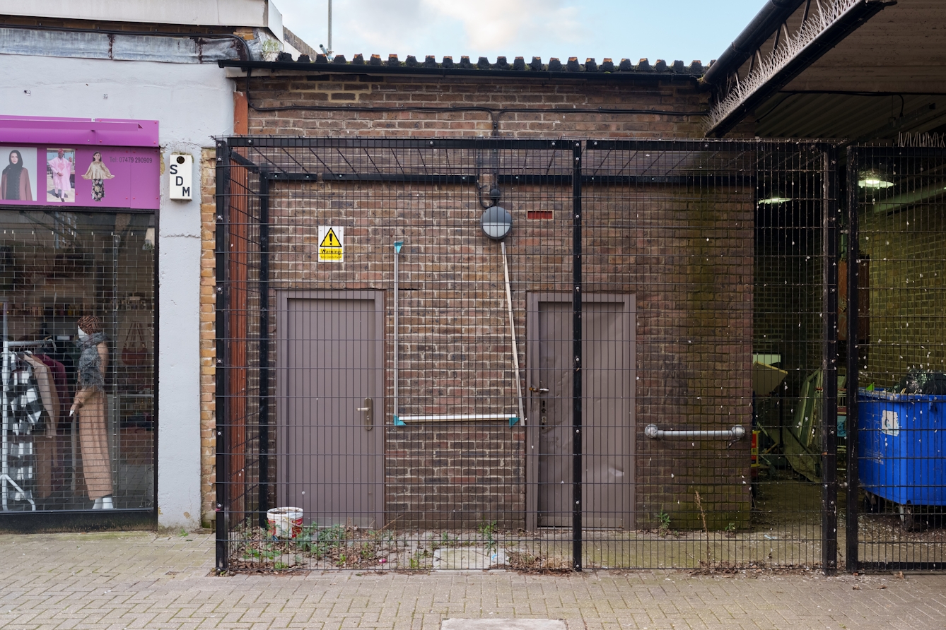 A photograph of two doors surrounded by a large metal cage. Inside the cage there are weeds and rubbish bins.