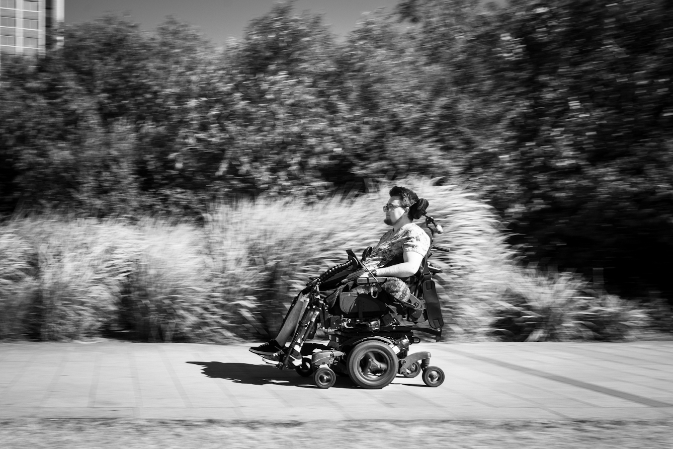 Black and white photograph of an individual racing along the pavement in their electric wheelchair.