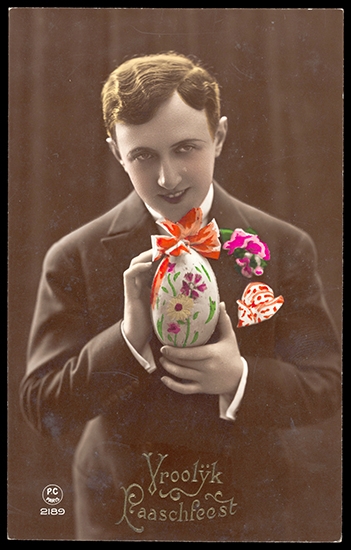 Young man holding decorated ester egg, with brightly coloured flowers in his lapel and a handkerchief in his breast pocket. The photograph is hand colourised.