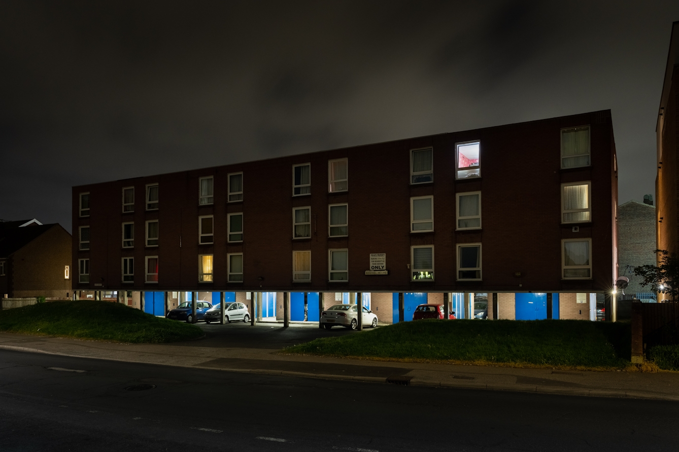 A photograph of a block of flats at night. There are cars parked under the structure, illuminated by security lights, with a series of bright blue doors visible. There are lights on and shining through the curtains of some of the windows, while others remain in darkness.