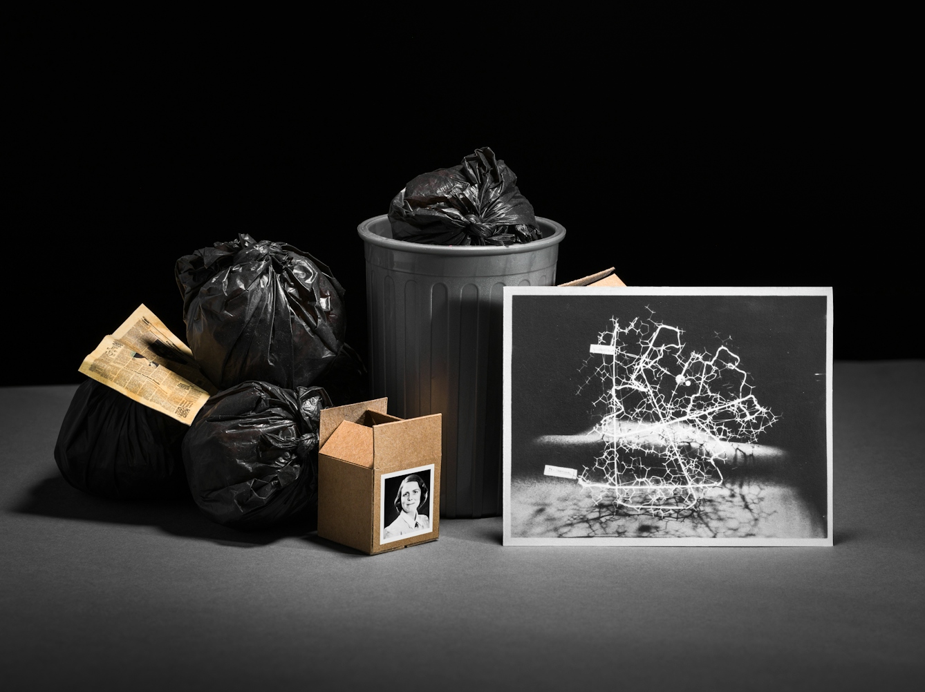 Photograph of a set-built scene made up of a grey card horizontal base against a black vertical background. In the centre of the image is a grey dustbin containing black bin bags. Surrounding the bin are more black bags and brown cardboard boxes. Leaning against one of the cardboard boxes is a black and white photographic print of a protein model.