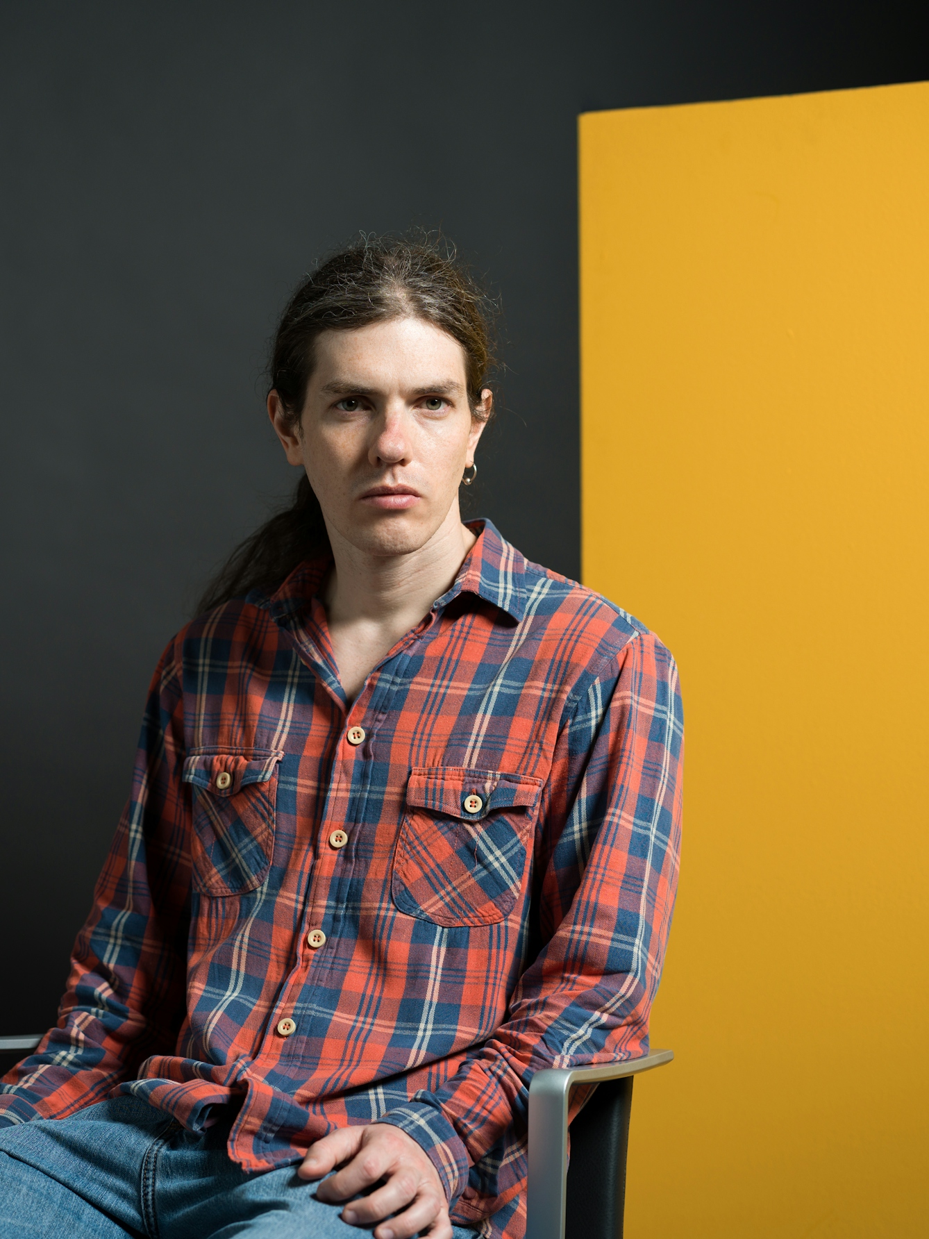 Colour photograph half-length portrait of Sam Castell-Ward, who is seated in a chair. Sam is white and has a serious expression. Sam has brown hair swept back into a ponytail and a single hoop earring in their left ear. Sam wears a red and blue plaid button-down shirt and jeans. 
