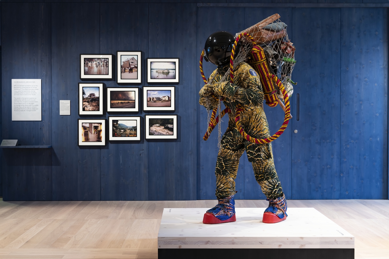 A photograph showing a life-size figure standing on a low wooden platform mounted on a black plinth beyond which is a dark blue painted wooden  panelled wall on which exhibits hang. The life-size figure wears: blue and red cotton patterned moon boots with red rubber soles; a green and yellow patterned cotton space suit and gloves; and a reflective black spherical helmet. The refugee astronaut carries a net on his or her back filled with possessions such as a suitcase, a book, a teapot and a telescope. On his or her back, there are also breathing tanks with long tubes connected to the spacesuit, made from red and yellow patterned cotton. 