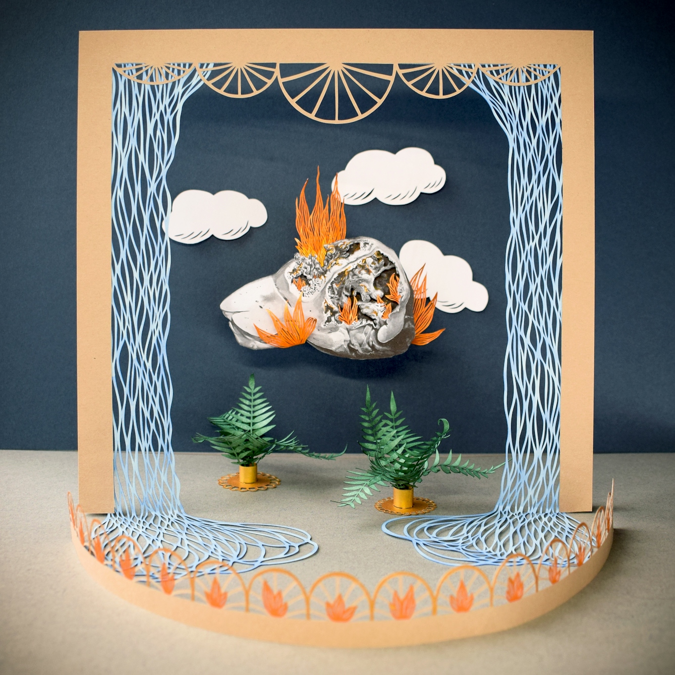Colourful papercut artwork with many separate elements on different levels creating a scene with depth, perspective and shadows. The scene shows a theatrical looking orange toned set with a stage front and curtain-like frame. Within stage there are 2 fern plants in pots on the floor and hanging from the blue backdrop are white clouds and  a monotone drawing of the human liver which appears to have flames emanating from it.