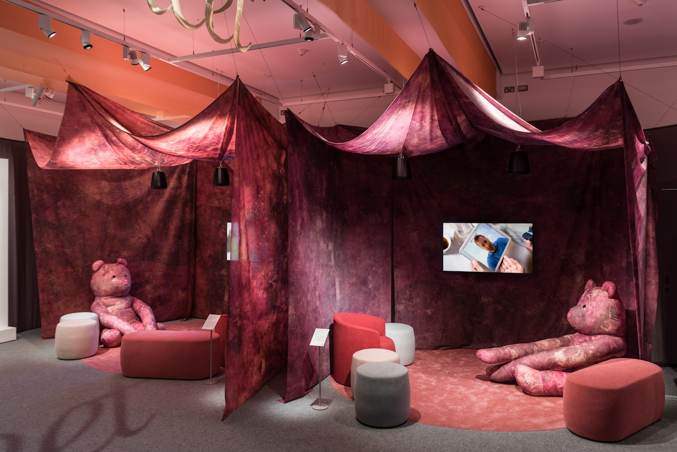 Photograph of a section of a gallery exhibition, showing two large purple and pink tie-dyed 'tents'. Inside each tent is a video screen showing a film. Each tent contains a large purple and pink tie-dyed teddy bear with long arms surrounded by several seats.