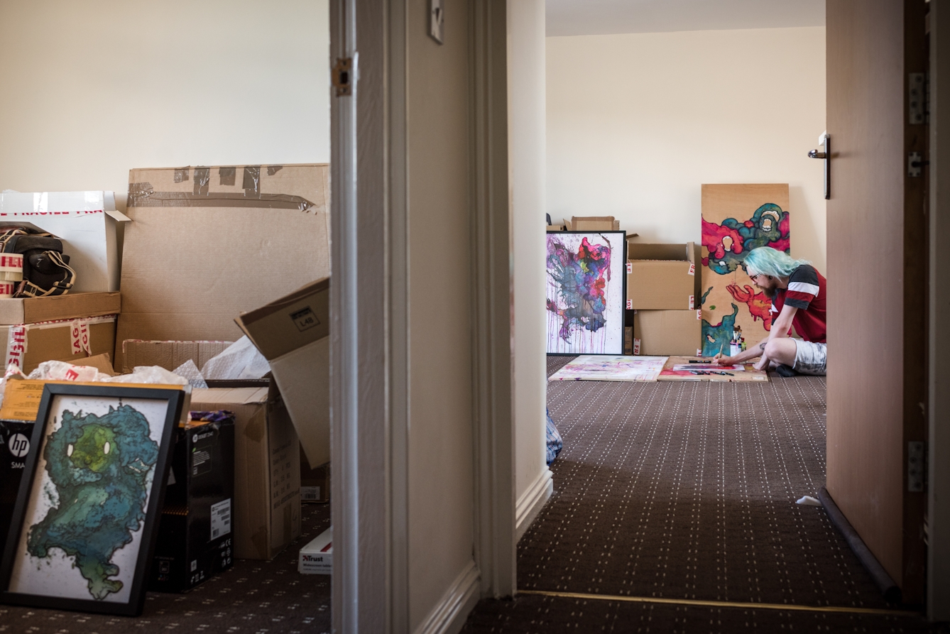 Photograph down the corridor of a flat showing unpacked boxes in one room and a man sitting on the floor creating a colour artwork in another.
