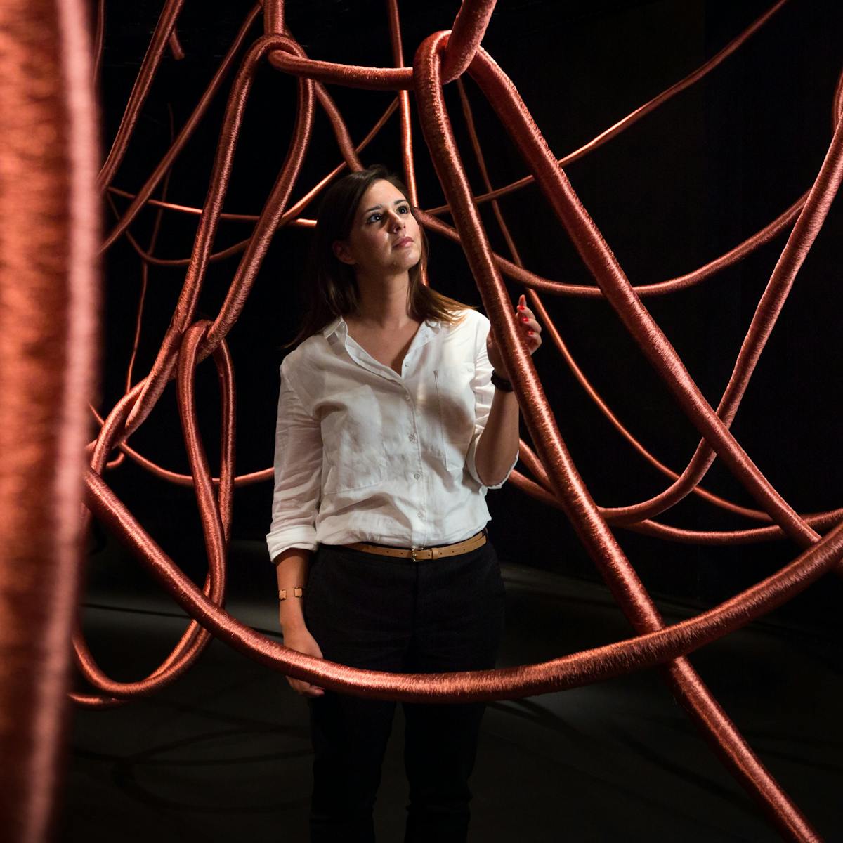 Photograph of a young woman exploring the exhibition, Alice Anderson: Memory Movement Memory Objects. She is standing in a dark gallery space surrounded by think hanging copper ropes.