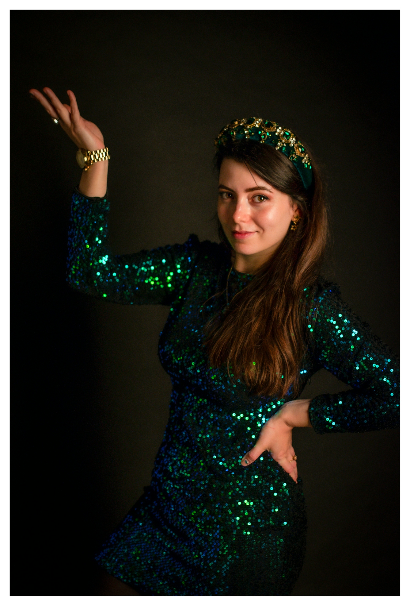 Photographic portrait of a white woman wearing a sequinned green and blue dress with matching hairband, stood against a black background. She is looking to camera with an quiet smile, one hand on her hip, the other raised in the air, palm open upwards.