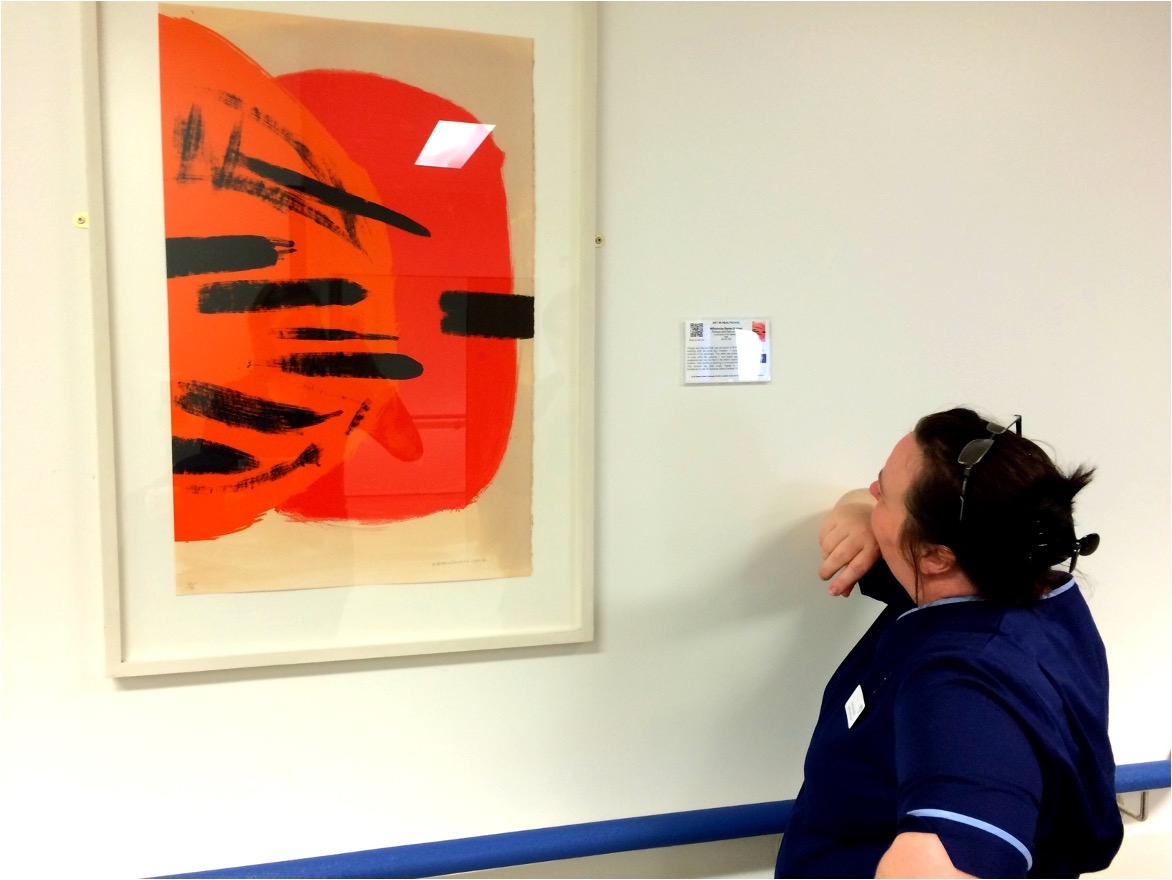 Colour photograph showing a woman looking closely at a framed painting on a wall. She is wearing a blue nurse's uniform. The picture is an abstract one featuring red circles and thick black brushstroke lines.