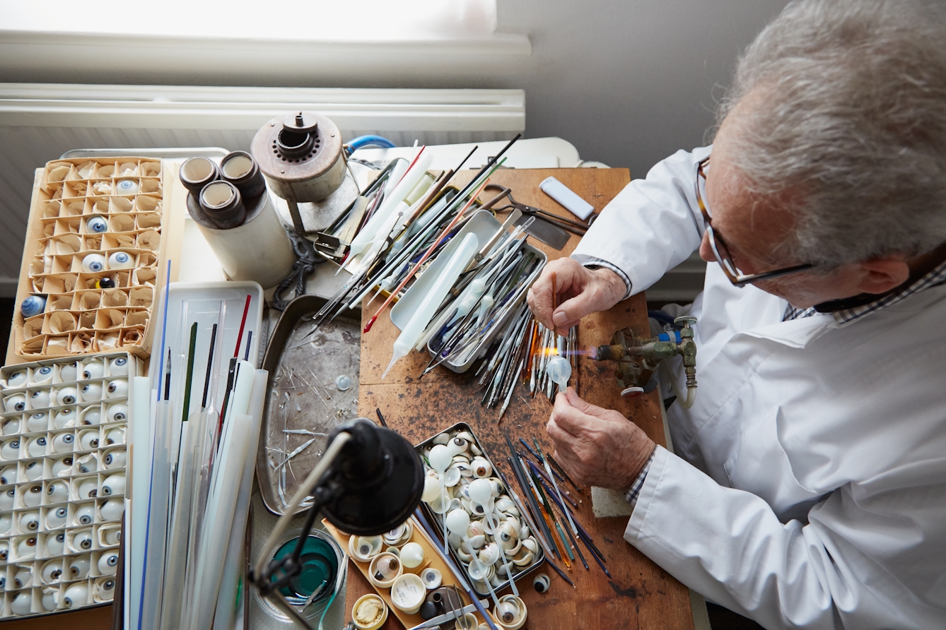 Photograph from above looking down on a man working at a workbench. He is holding a glass tube over a gas torch. On the surface of the workbench he is surrounded by more glass tubing, thin tools and trays containing glass eyes.