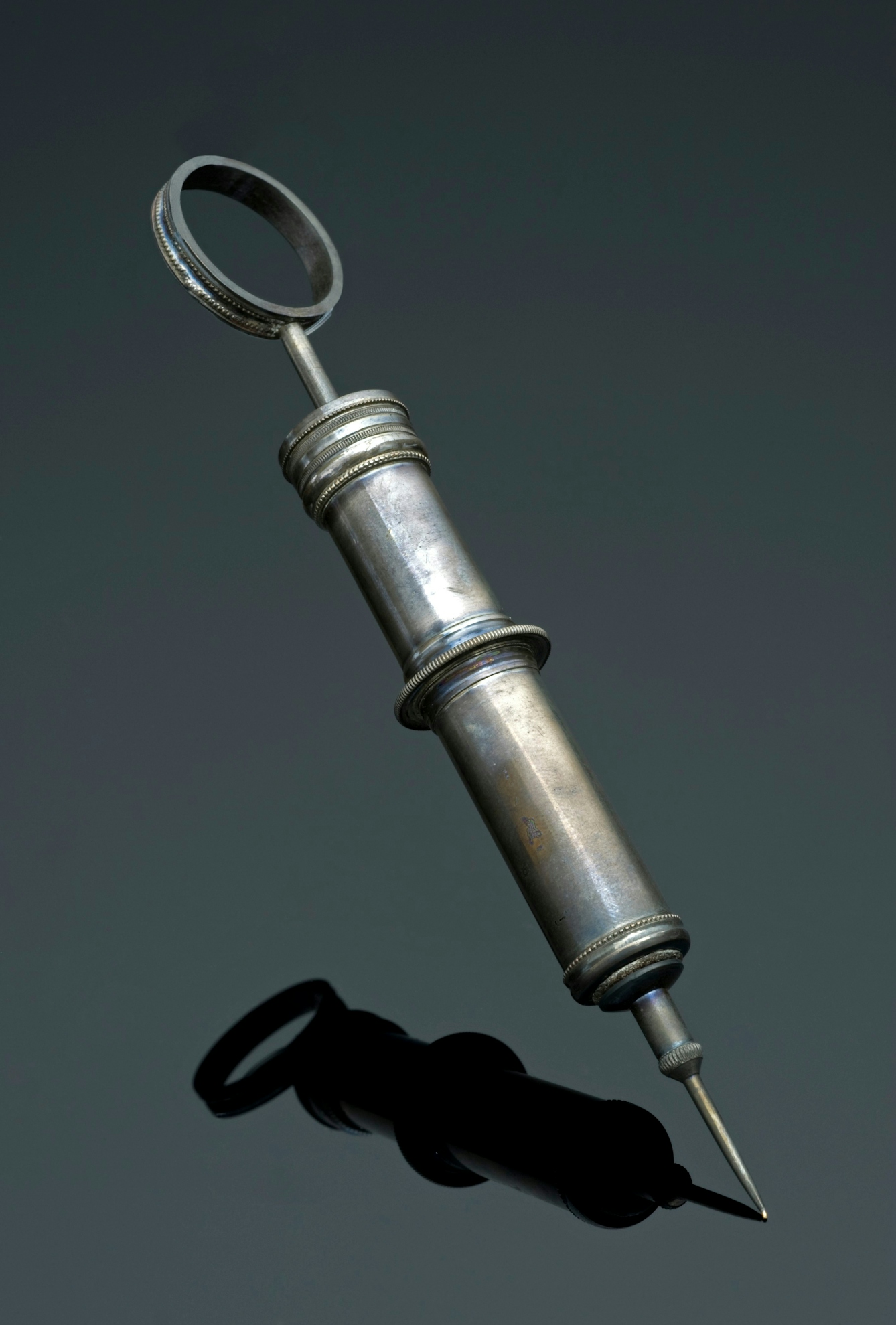Syringe made of metal (with a slight patina) with a large circular plunger at the end and a thick needle.