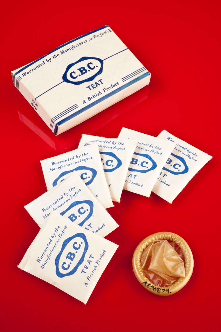 Photo of small square white card boxes with blue writing, neatly displayed next to a condom. The background is red.
