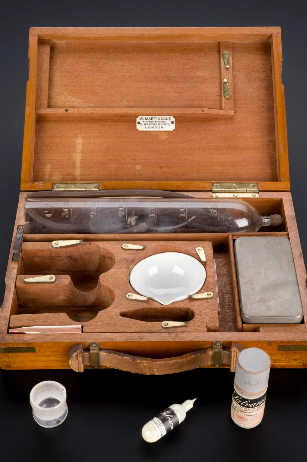 An open wooden box containing a Salvarsan kit, including Salvarsan in a glass phial, hypochloric acid and litmus paper, plus other apparatus to administer the drug.
