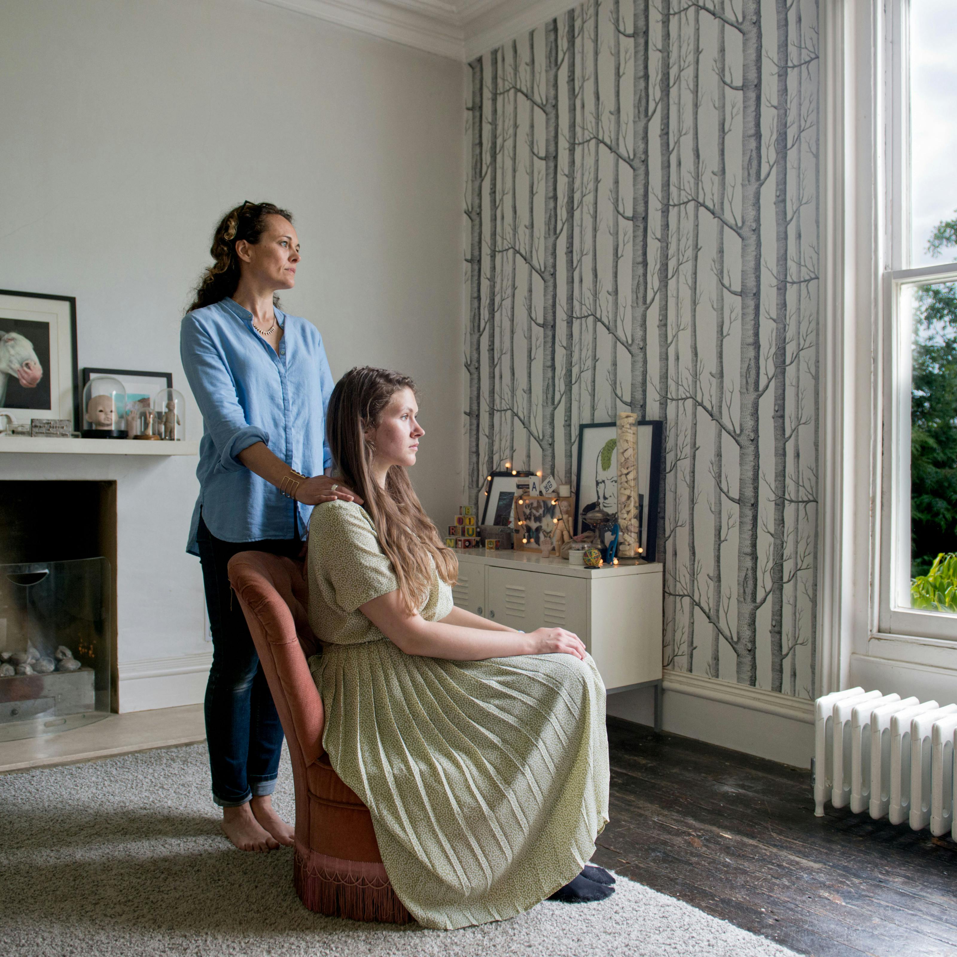 Photograph of a young woman and a mother figure in a domestic living room. The young woman is seated in a long dress and and the mother figure stands behind her, hands on her shoulders. They are both gazing out of the window.