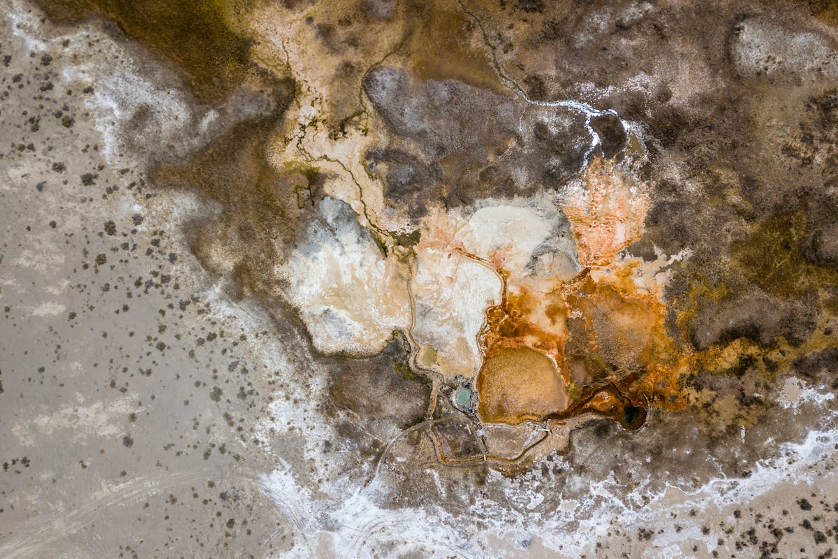 Aerial colour photograph taken from 1,500 metres above ground level, looking down on a geothermal hot spring. The image is abstract in nature making it difficult to get a sense of scale. The overall tones are a brown, red, rust colour with patches of white. There is a small area of green liquid surrounded by a patchwork of alluvial features. The landscape is Barron and lacking in vegetation. Small snaking lines can just be made out which could be the tracks of unseen vehicles.